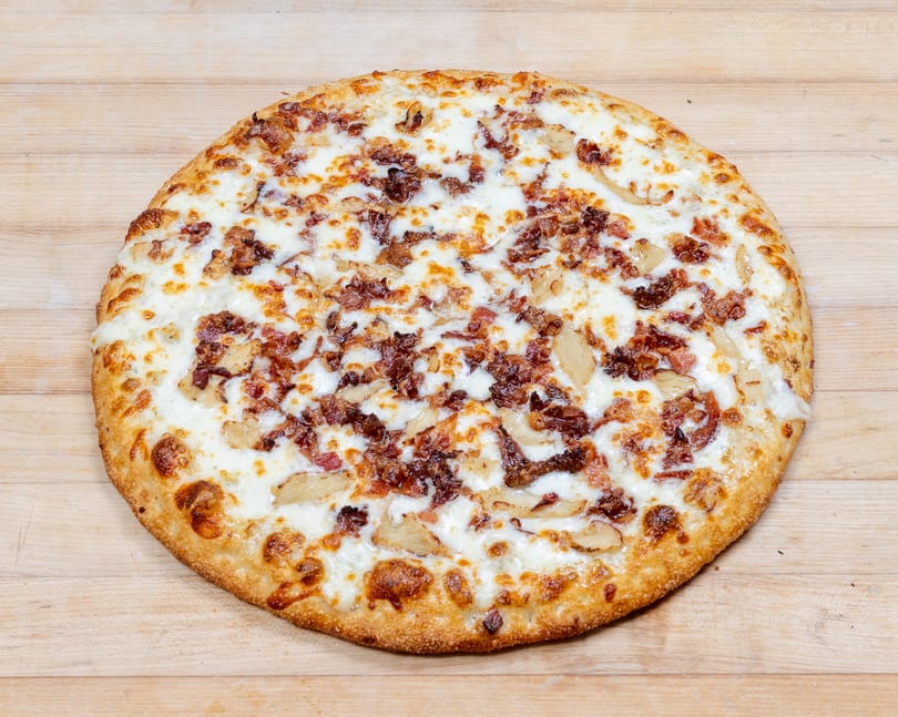 Round pizza with white sauce, melted cheese, bacon, and chicken on a wooden surface.