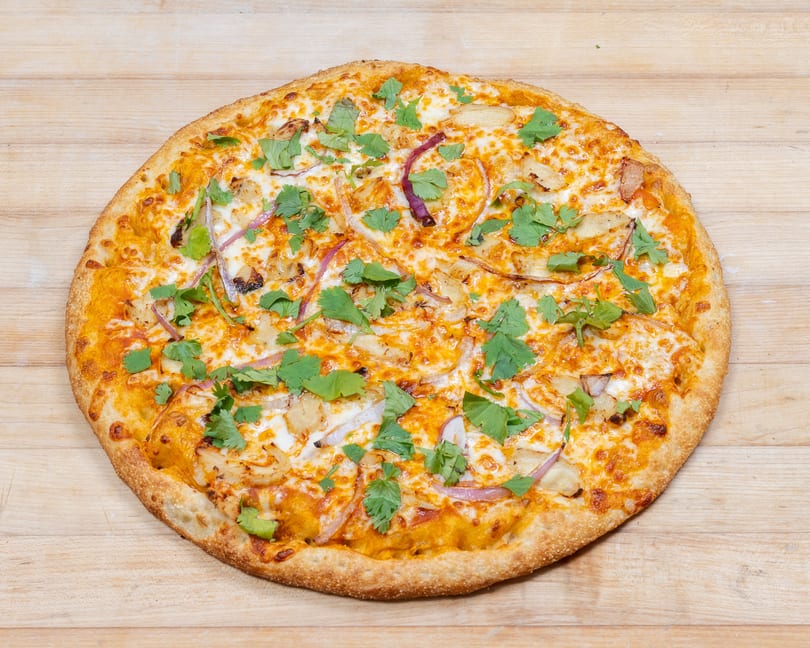 Whole chicken tikka pizza with red onions, cilantro, and cheese on a wooden surface.
