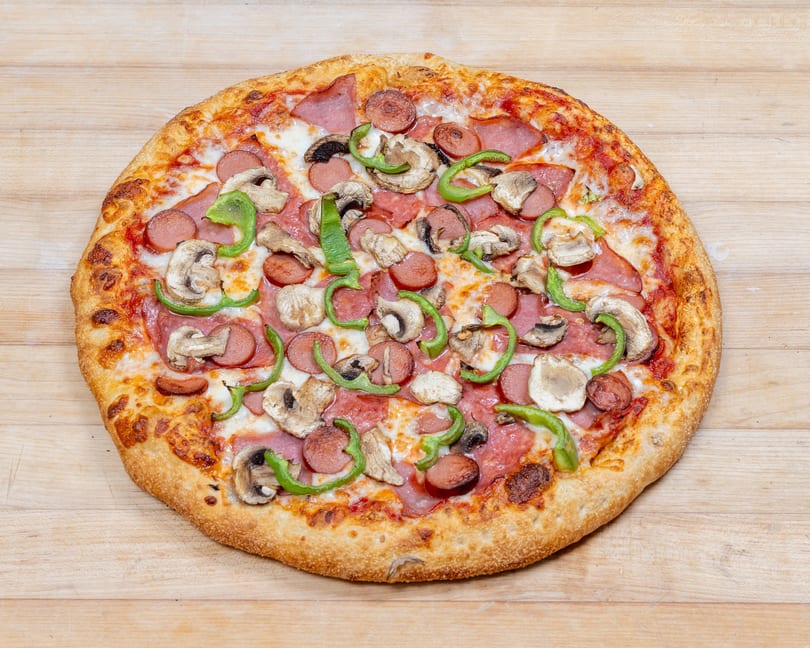 A whole pizza with sausage, ham, mushrooms, and green bell peppers on a wooden surface.