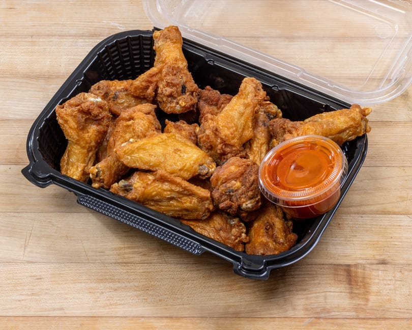 Crispy fried chicken wings in a black takeout container with a side of orange dipping sauce.