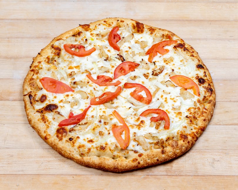 A whole pizza with white sauce, chicken, onions, and tomato slices on a wooden surface.