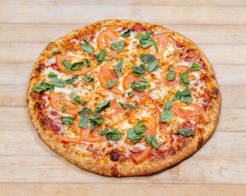 A whole Margherita pizza with sliced tomatoes and fresh basil on a wooden surface.