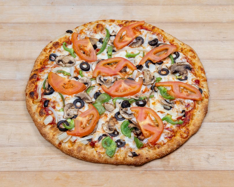 A whole vegetarian pizza with tomatoes, mushrooms, olives, onions, and green peppers on a wooden surface.