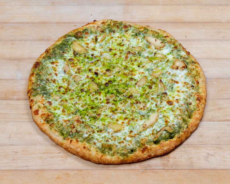 A whole pesto chicken pizza with melted cheese and a golden crust on a wooden surface.