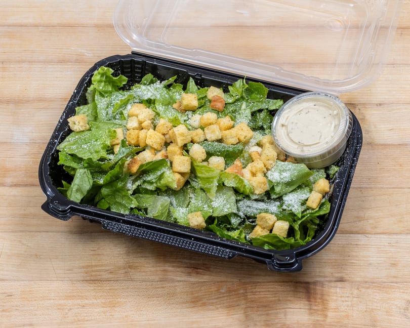 Caesar salad with croutons and dressing in a black plastic container on a wooden surface.