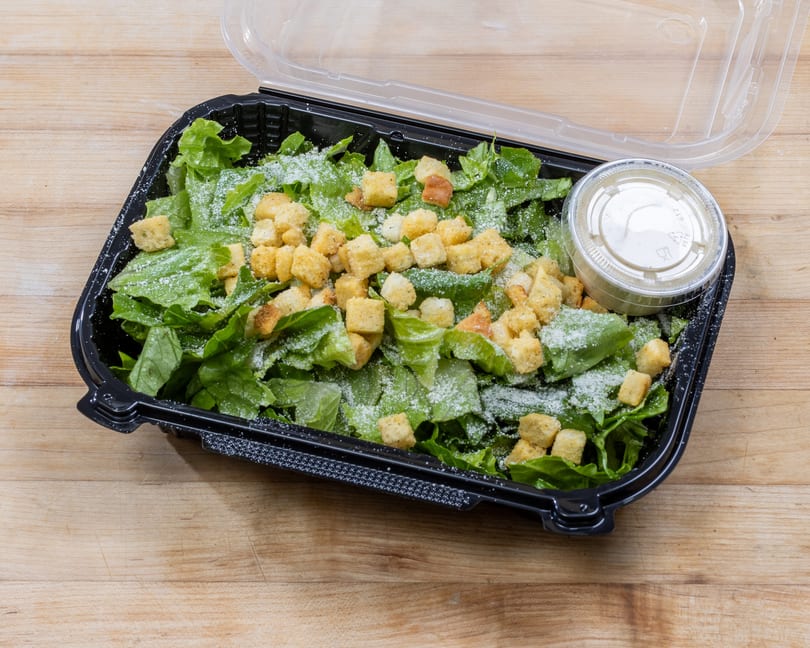 Caesar salad with croutons and dressing in a black plastic takeout container on a wooden surface.