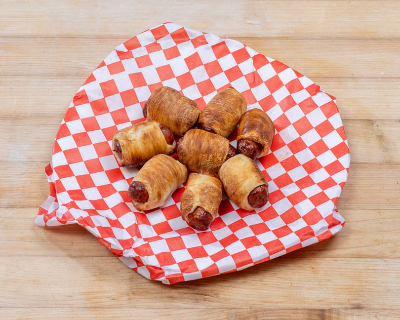 Seven pigs in a blanket, wrapped in golden pastry, on red and white checkered paper.