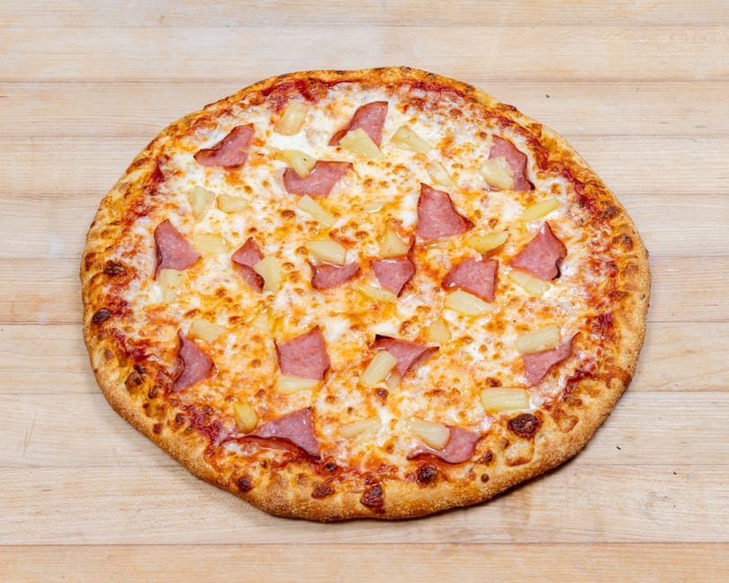 A whole Hawaiian pizza with ham and pineapple on a light wooden surface.